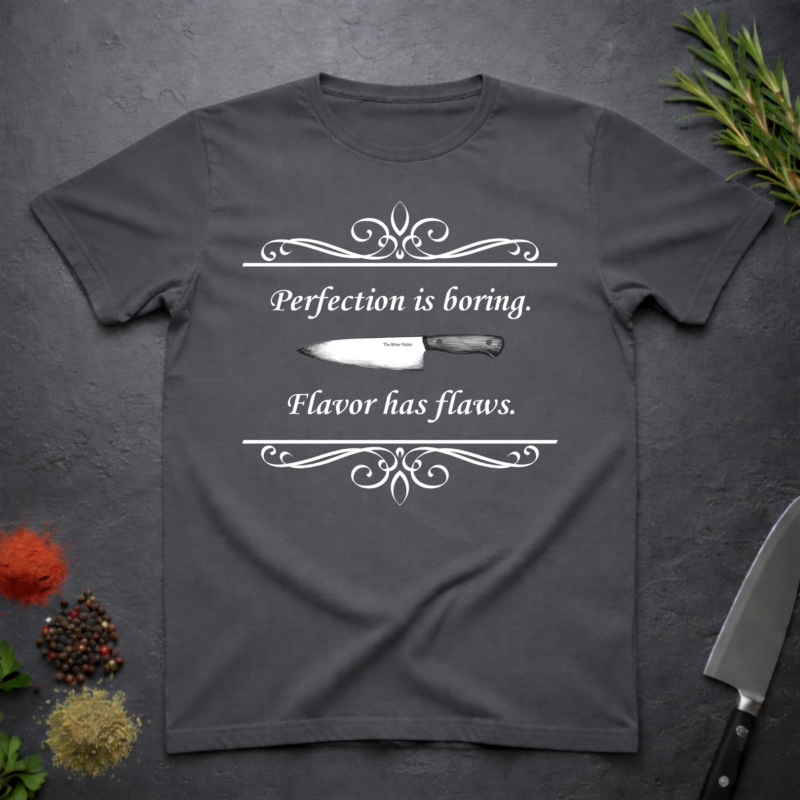 Charcoal grey t-shirt with a knife graphic and text "Perfection is boring. Flavor has flaws." On a dark surface. 