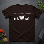 Chocolate Brown t-shirt with text 'Life Cycle of a Chicken' and graphic of an egg, chick, hen, frying pan on a dark surface.