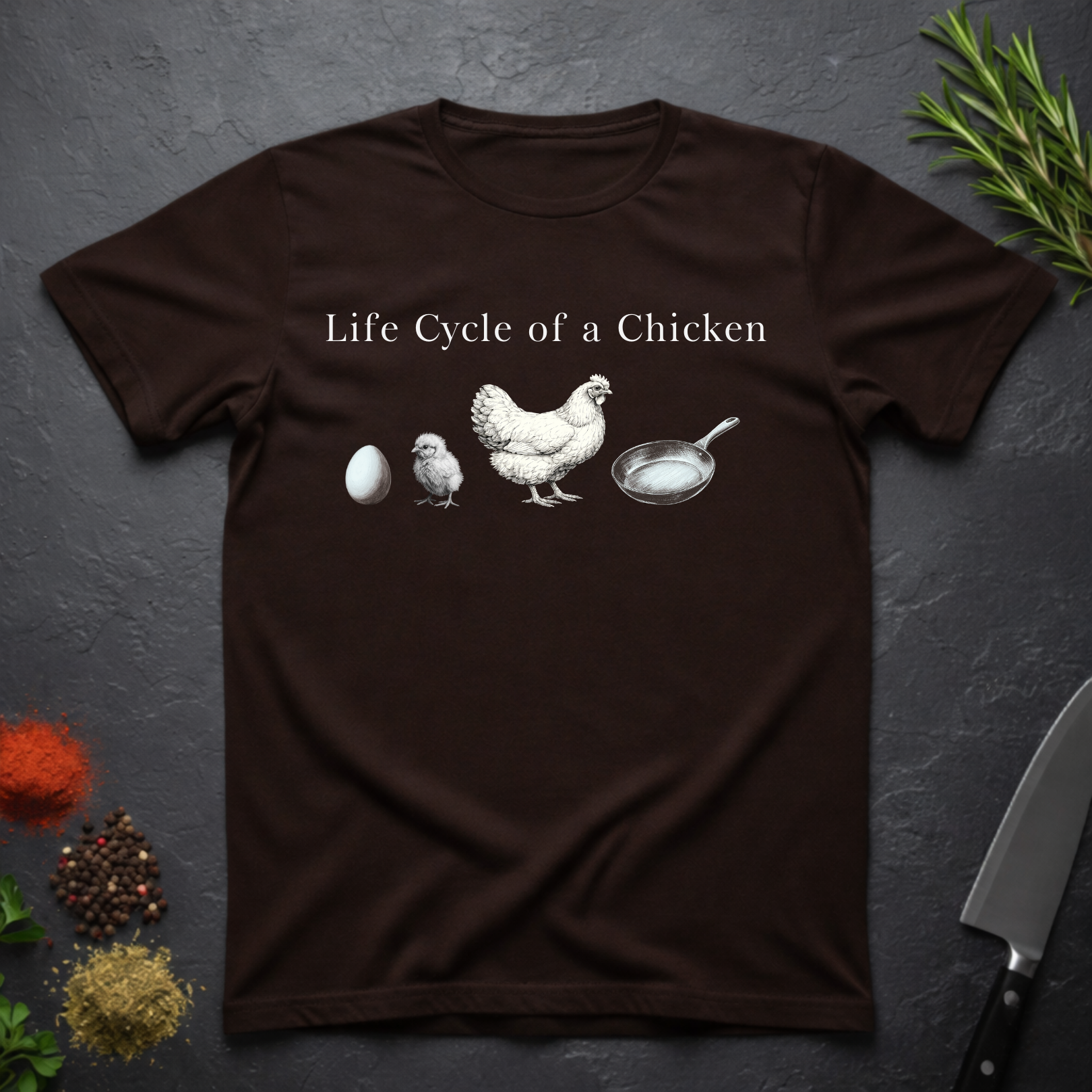 Chocolate Brown t-shirt with text 'Life Cycle of a Chicken' and graphic of an egg, chick, hen, frying pan on a dark surface.