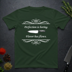 Forest green t-shirt with a knife graphic and text "Perfection is boring. Flavor has flaws." On a dark surface. 