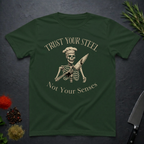 Forest green t-shirt with skeleton design and text "Trust your Steel, Not Your Senses" on a dark surface with decorative elements.
