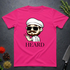 Heliconia pink t-shirt with a cartoon character chef and 'HEARD' text on a dark surface.