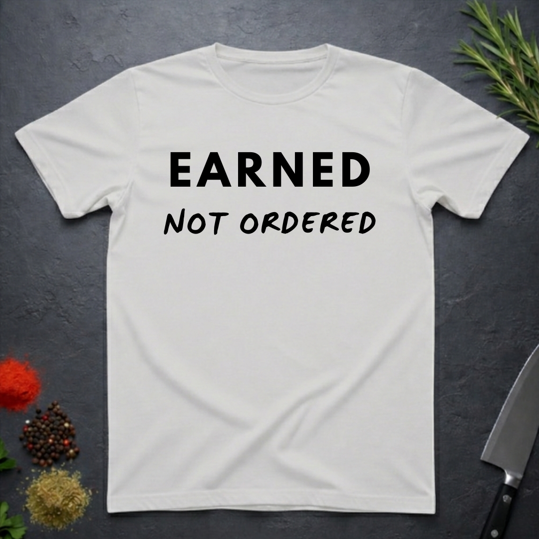 Ice Grey t-shirt with 'Earned Not Ordered' text on a dark surface with decorative elements.