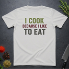 Ice Grey t-shirt with 'I cook because I like to eat' text on a dark surface with kitchen items.