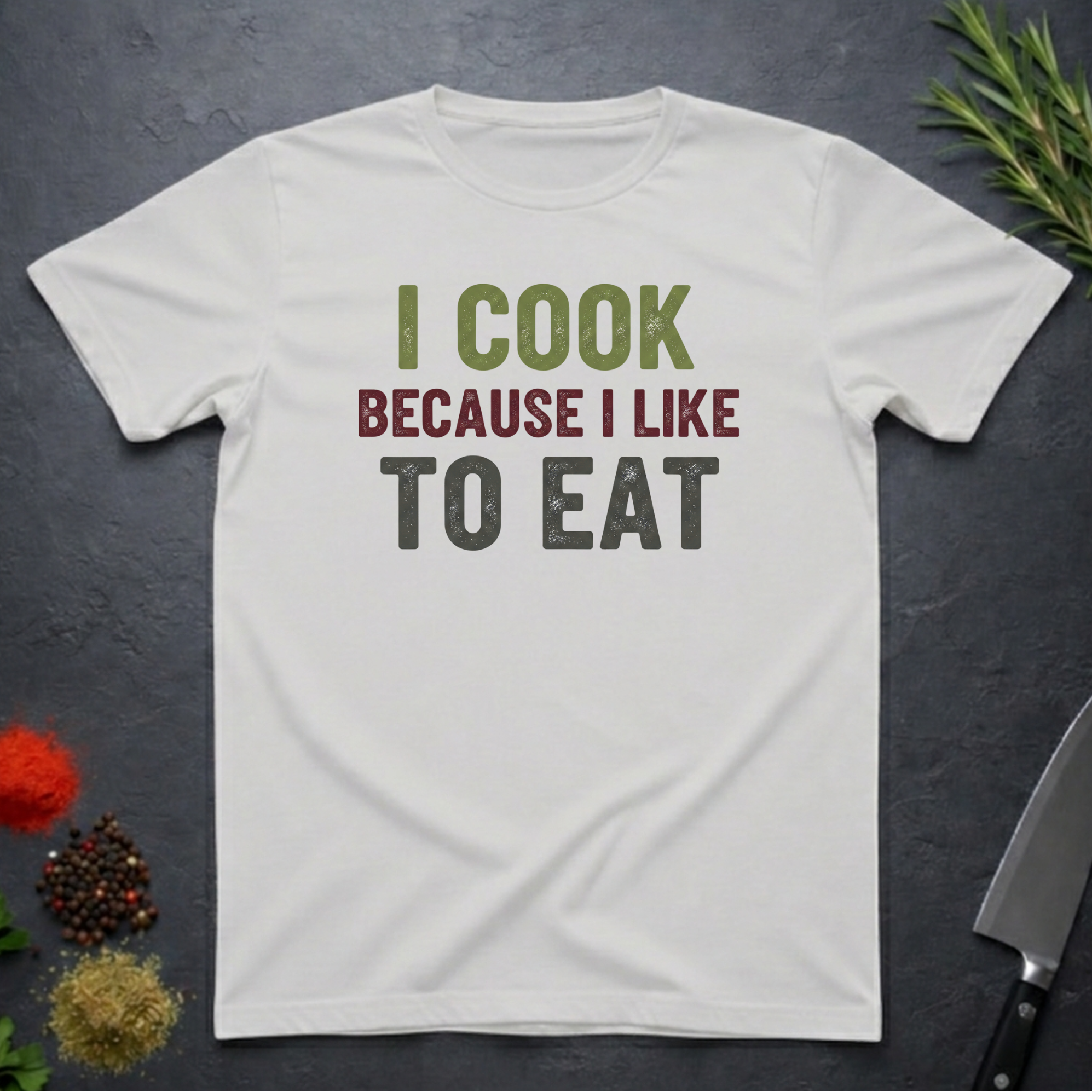 Ice Grey t-shirt with 'I cook because I like to eat' text on a dark surface with kitchen items.