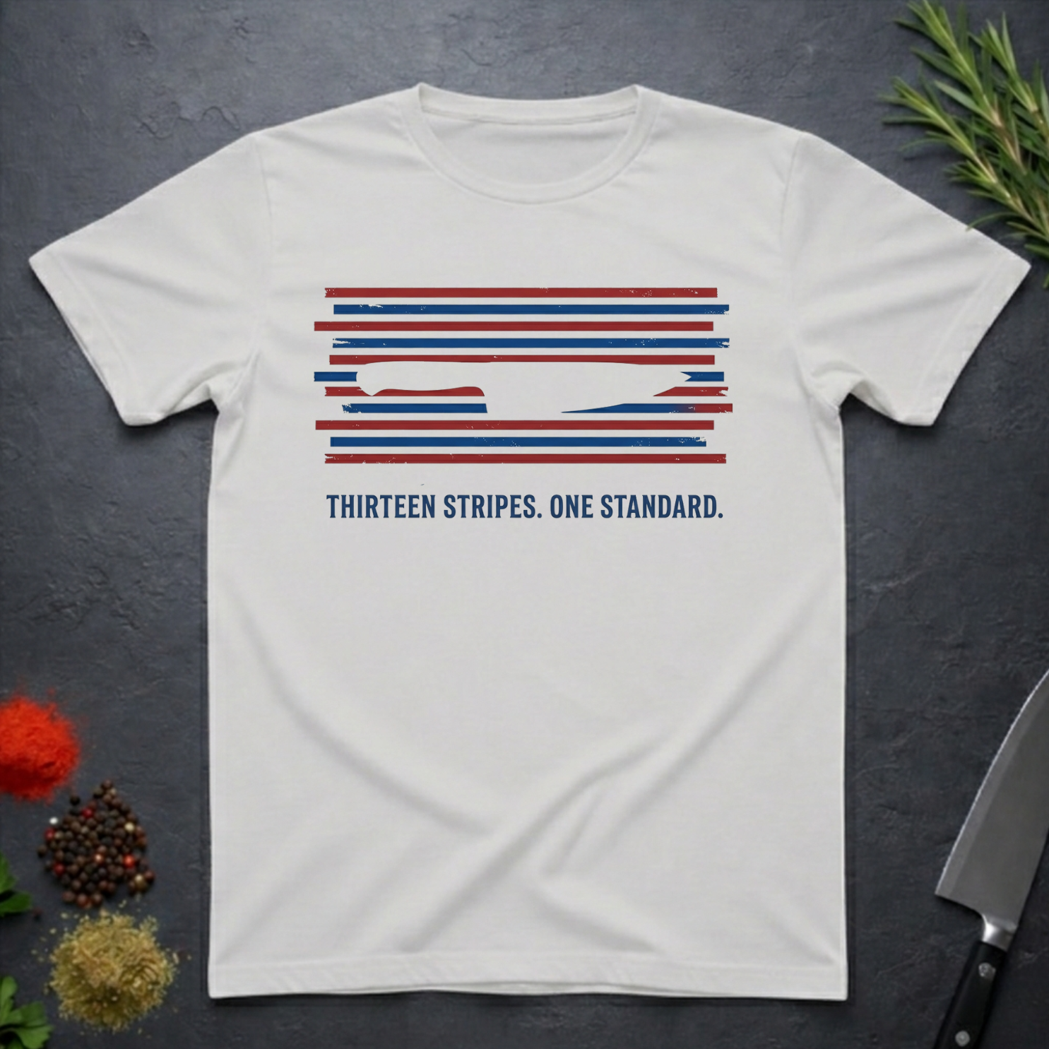 Ice grey t-shirt with 13 red, white, and blue stripes with a cut out (negative space) image of a chef's knife and text "THIRTEEN STRIPES. ONE STANDARD." on a dark surface.