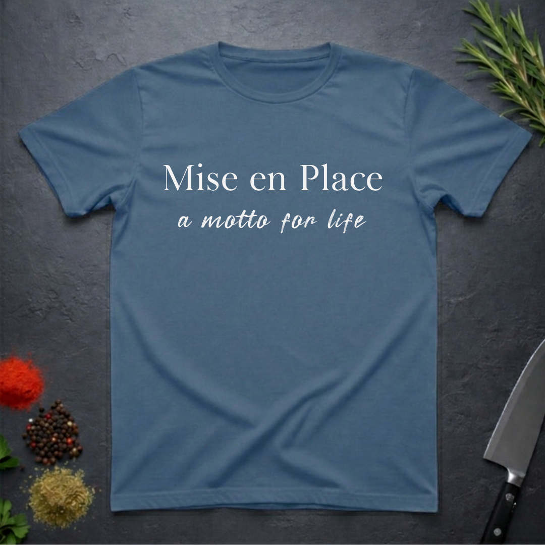 Indigo Blue t-shirt with 'Mise en Place a motto for life' text on a dark surface with kitchen items.