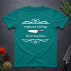 Jade dome colored t-shirt with a knife graphic and text "Perfection is boring. Flavor has flaws." On a dark surface. 