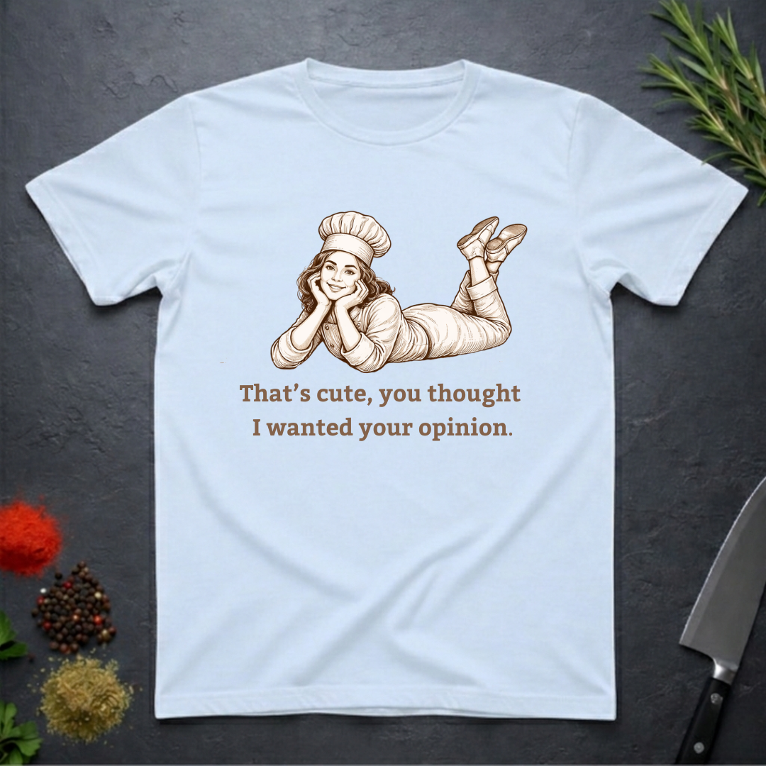 Light blue t-shirt with a graphic of a woman in a chef's outfit and text "That's cute, you thought I wanted your opinion." On a dark surface.