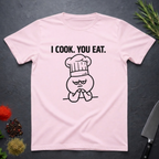 Light pink t-shirt with a graphic and text 'I COOK. YOU EAT.' on a dark surface.