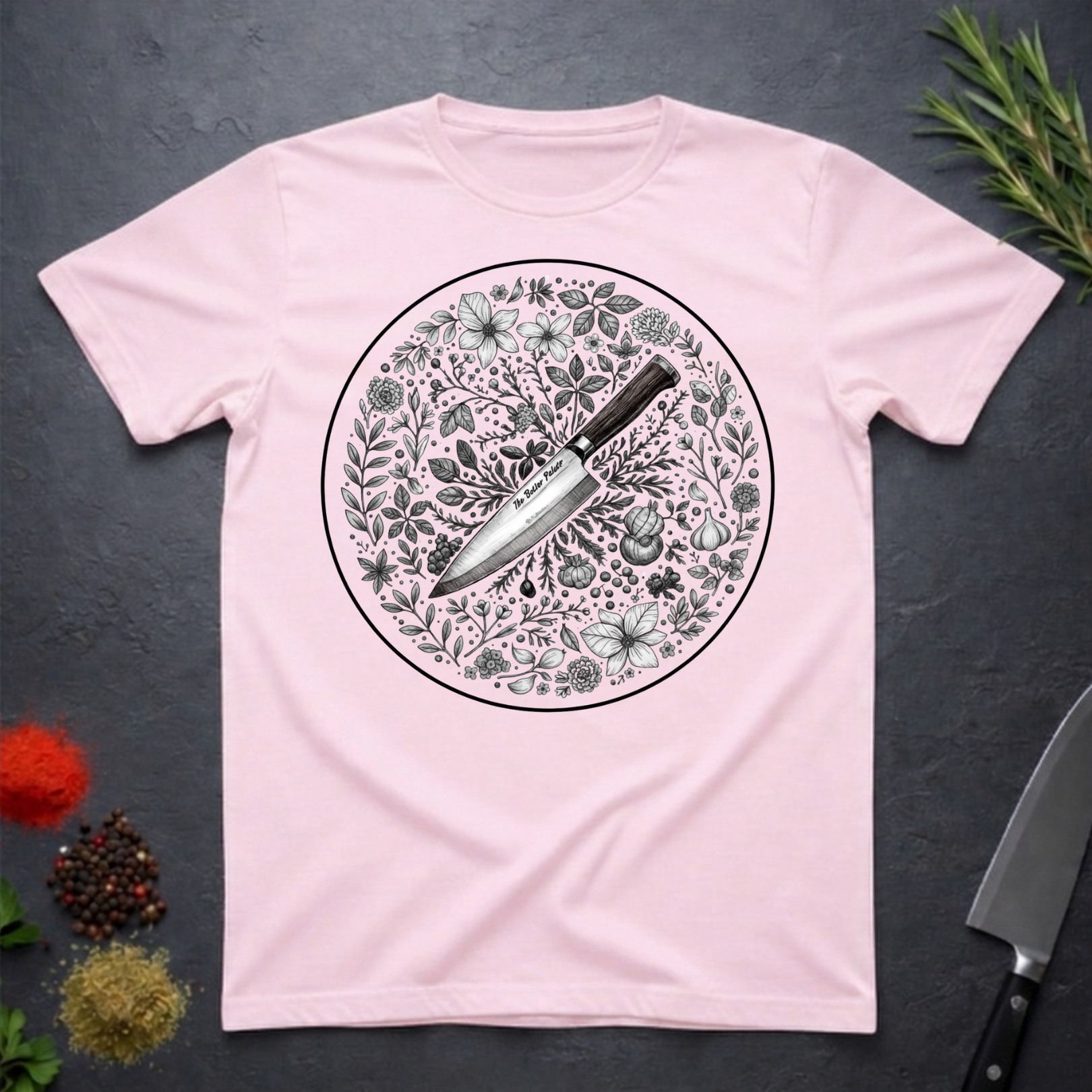 Light pink t-shirt with a circular floral and knife design on a dark surface.