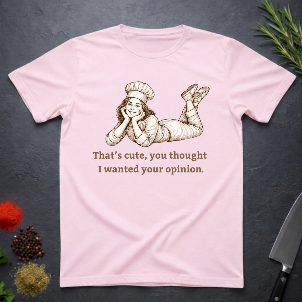 Light pink t-shirt with a graphic of a woman in a chef's outfit and text "That's cute, you thought I wanted your opinion." On a dark surface.