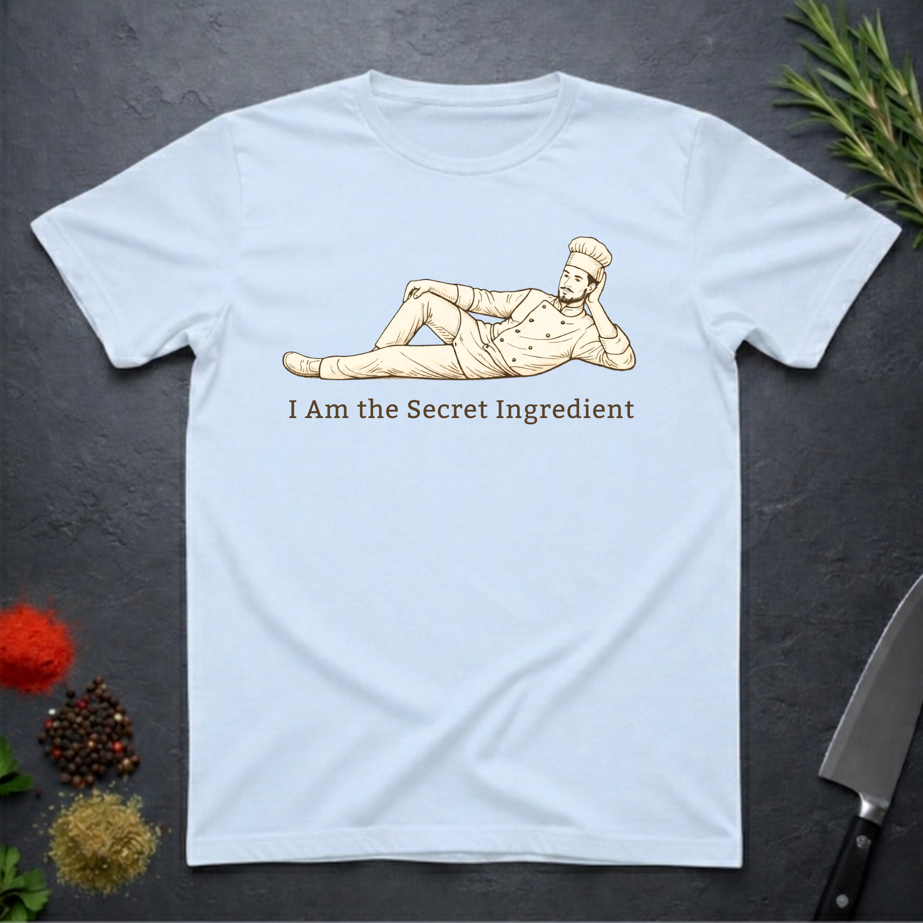 Light Blue t-shirt with a graphic of a chef lying on his side and text 'I Am the Secret Ingredient' on a dark surface with kitchen items.