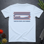 Light blue t-shirt with 13 red, white, and blue stripes with a cut out (negative space) image of a chef's knife and text "THIRTEEN STRIPES. ONE STANDARD." on a dark surface.