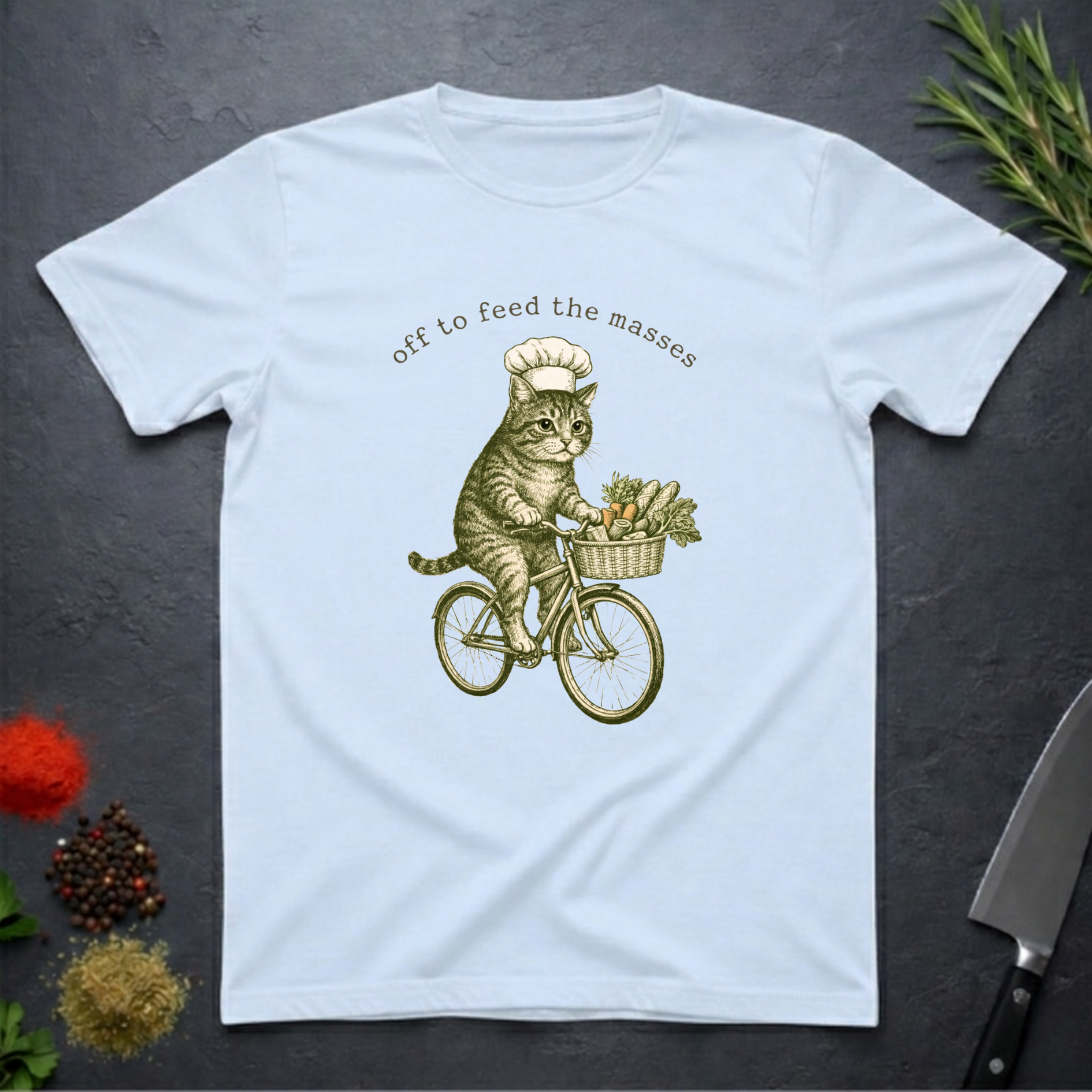 Light blue t-shirt with a graphic of a cat riding a bicycle and text 'off to feed the masses' on a dark surface.