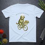 Light blue t-shirt with a graphic of a dog riding a bicycle with vegetables and text 'off to feed the masses' on a dark surface.