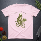 Light Pink t-shirt with a cat riding a bicycle graphic and text 'off to feed the masses' on a dark surface.