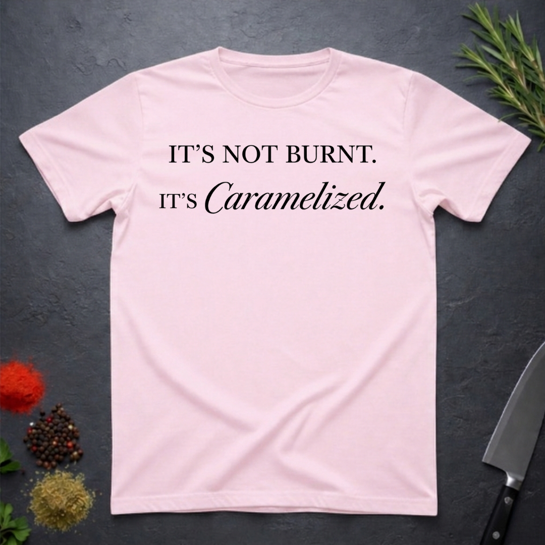 Light Pink t-shirt with text 'IT'S NOT BURNT. IT'S Caramelized.' on a dark surface with decorative elements.