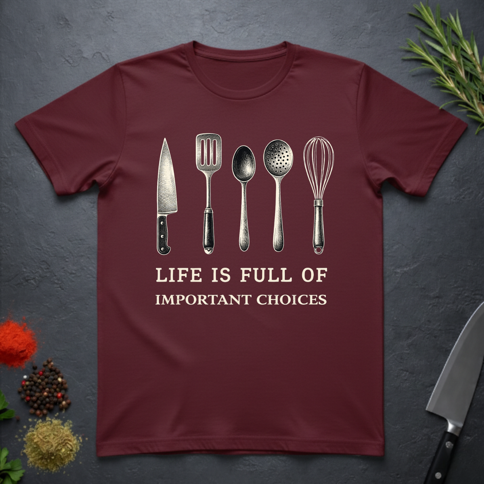 Maroon t-shirt with kitchen utensil graphics and text "Life is Full of Important Choices" on a dark surface.