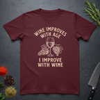 Maroon t-shirt with text "Wine Improves With Age. I Improve With Wine" in an off-white bold font. Image of wine glass and grape bunches separate text. From the Bitter Palate brand.