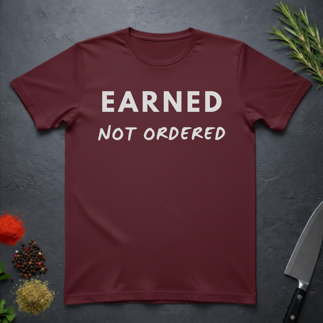 Maroon t-shirt with 'Earned Not Ordered' text on a dark surface with decorative elements.