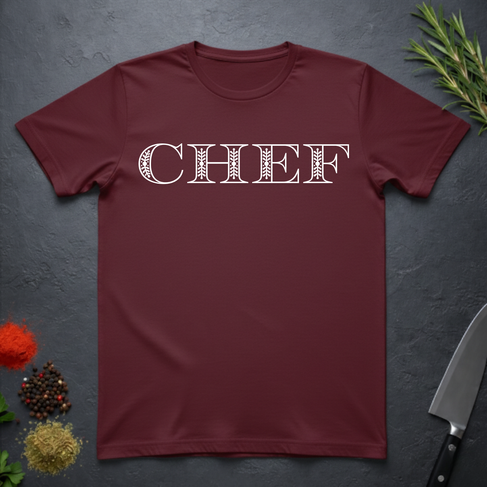 Maroon t-shirt with 'CHEF' printed in a decorative serif text on a dark surface with kitchen items.