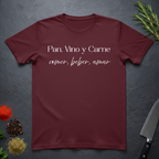 Maroon t-shirt with Spanish text "Pan, Vino y Carne" on a dark surface with decorative elements. 