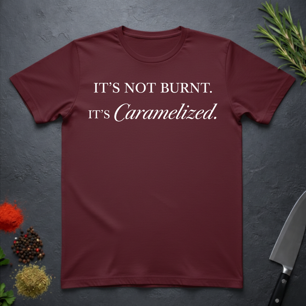Maroon t-shirt with text 'IT'S NOT BURNT. IT'S Caramelized.' on a dark surface with decorative elements.