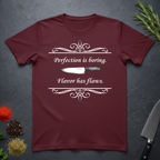 Maroon t-shirt with a knife graphic and text "Perfection is boring. Flavor has flaws." On a dark surface. 