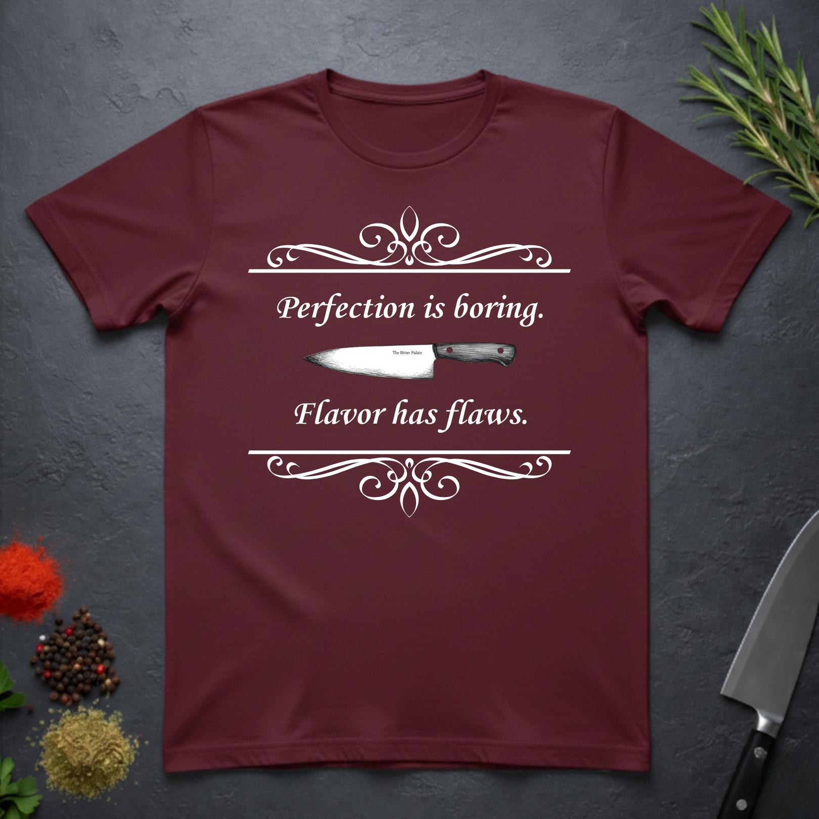Maroon t-shirt with a knife graphic and text "Perfection is boring. Flavor has flaws." On a dark surface. 