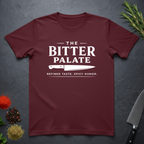 Maroon t-shirt with 'The Bitter Palate' logo on a dark surface with kitchen items.