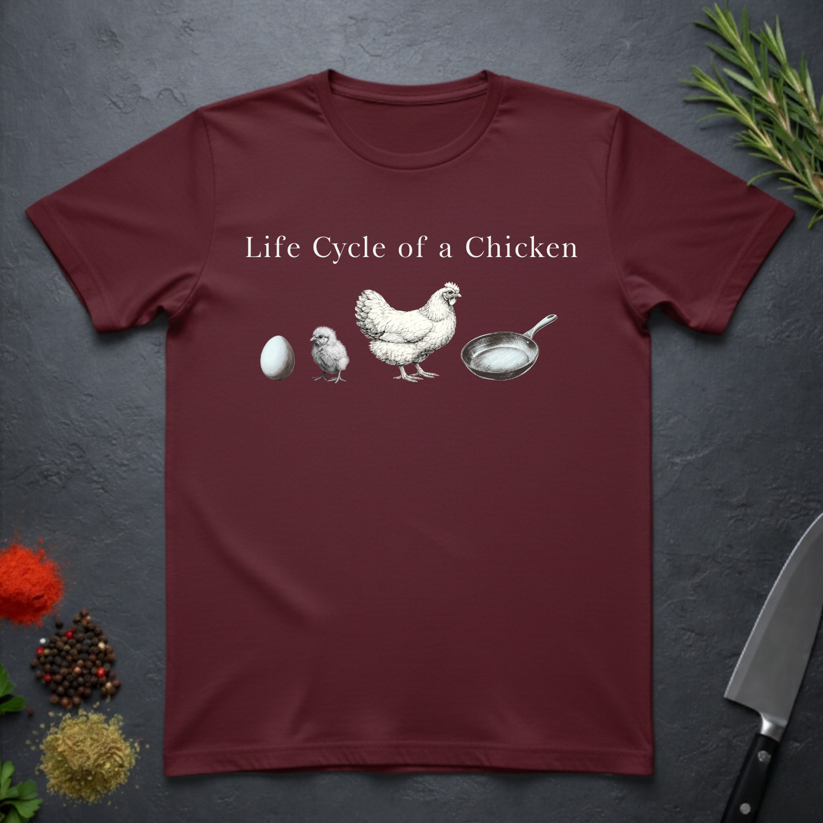 Maroon t-shirt with text 'Life Cycle of a Chicken' and graphic of an egg, chick, hen, frying pan on a dark surface.