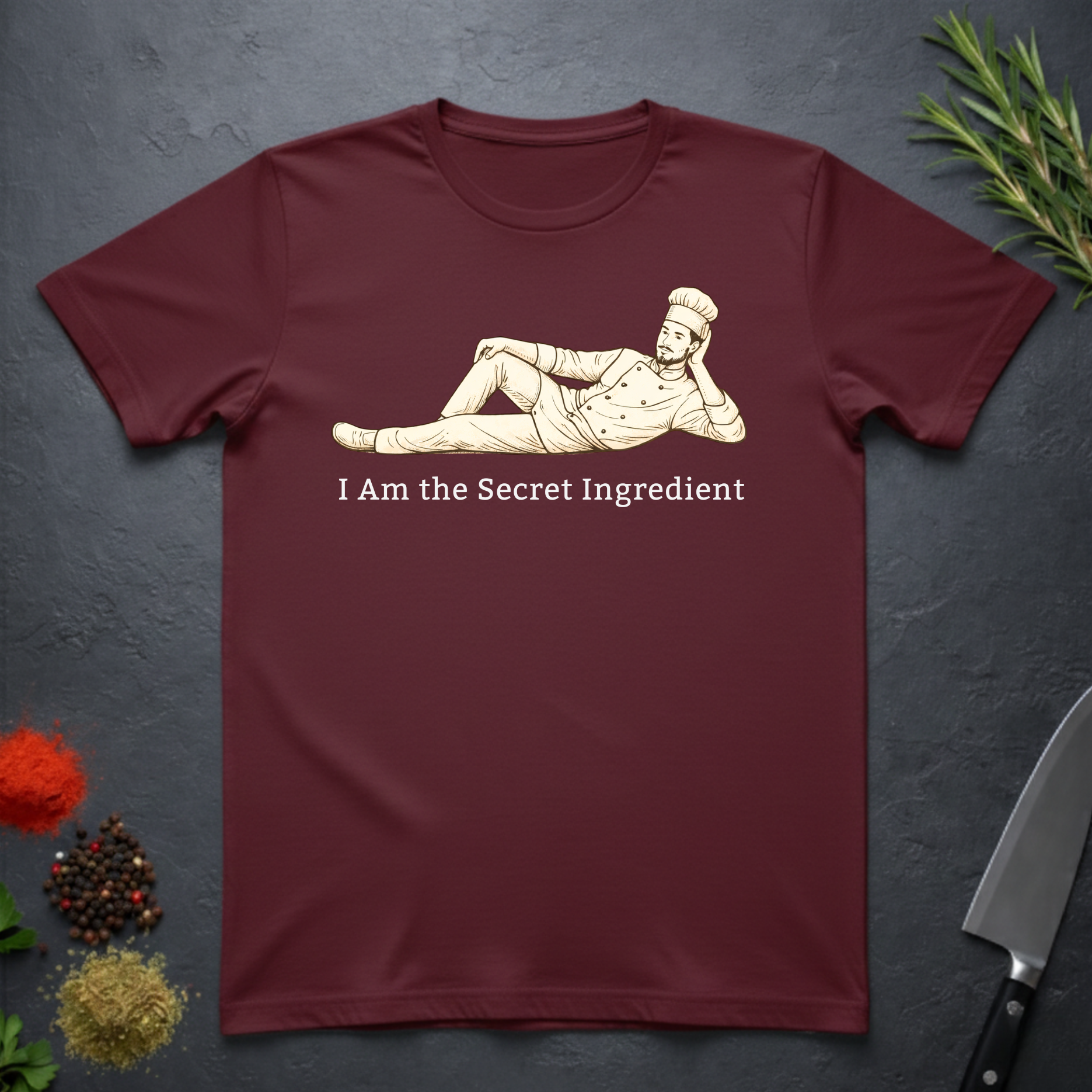 Maroon t-shirt with a graphic of a chef lying on his side and text 'I Am the Secret Ingredient' on a dark surface with kitchen items.