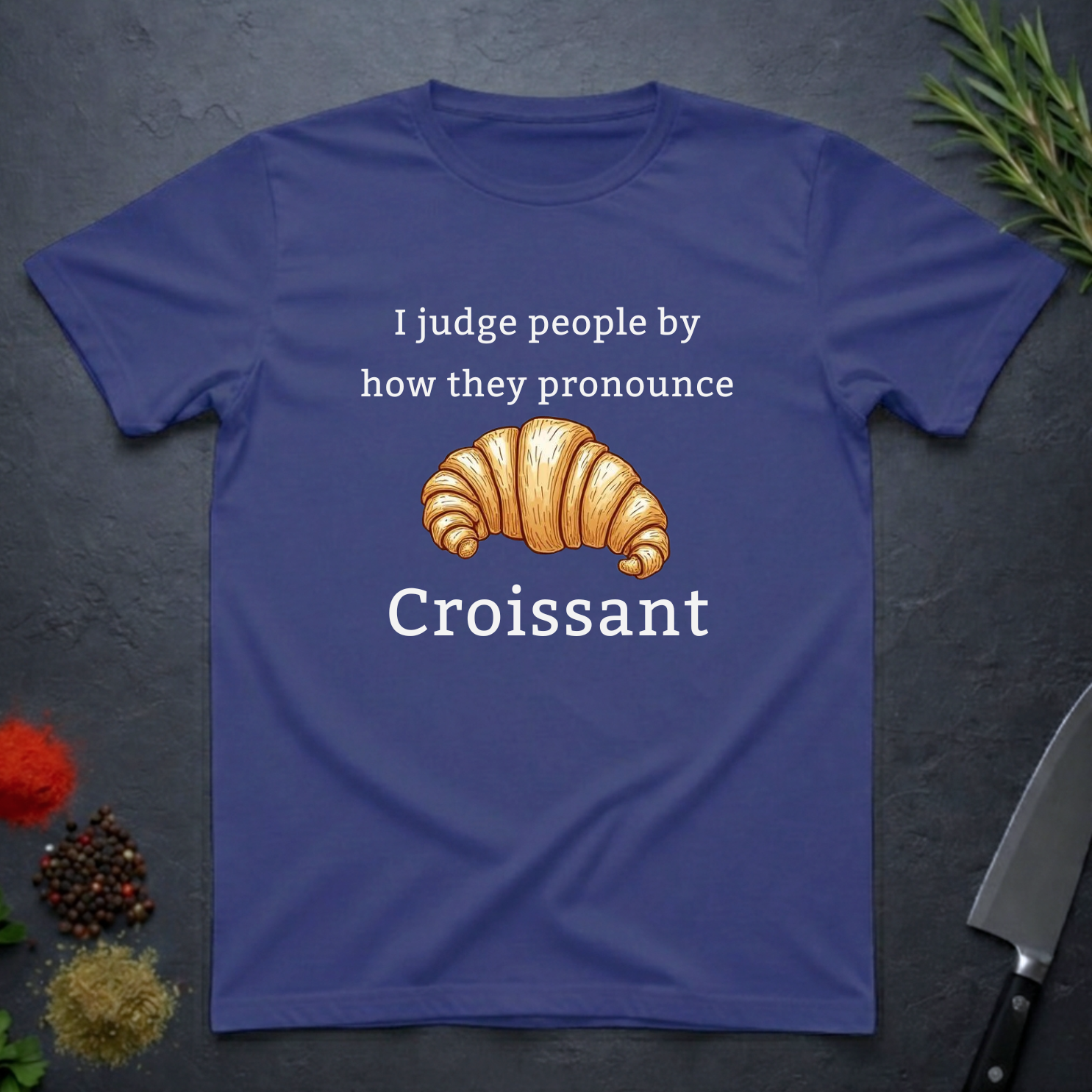 Metro blue t-shirt with a croissant graphic and text "I judge people by ho they pronounce Croissant" on a dark surface.