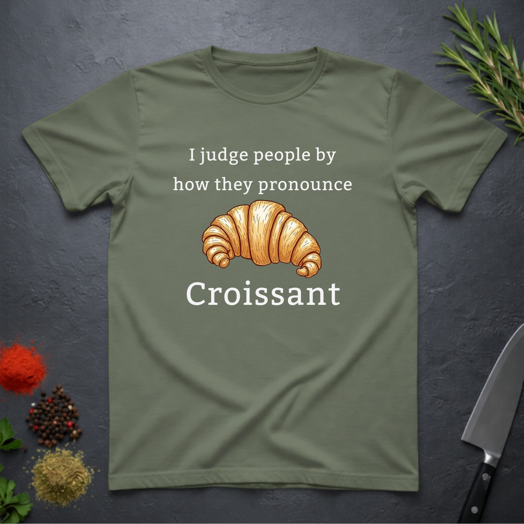 Sage green t-shirt with a croissant graphic and text "I judge people by ho they pronounce Croissant" on a dark surface.