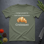 Sage green t-shirt with a croissant graphic and text "I judge people by ho they pronounce Croissant" on a dark surface.