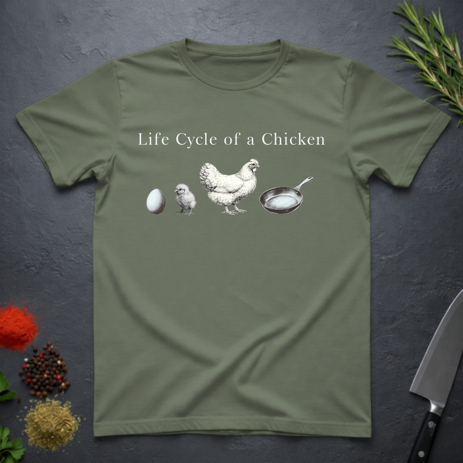 Military green t-shirt with text 'Life Cycle of a Chicken' and graphic of an egg, chick, hen, frying pan on a dark surface.