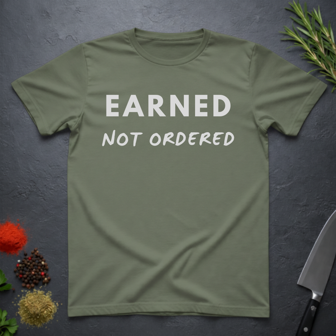 Military Green t-shirt with 'Earned Not Ordered' text on a dark surface with kitchen items.