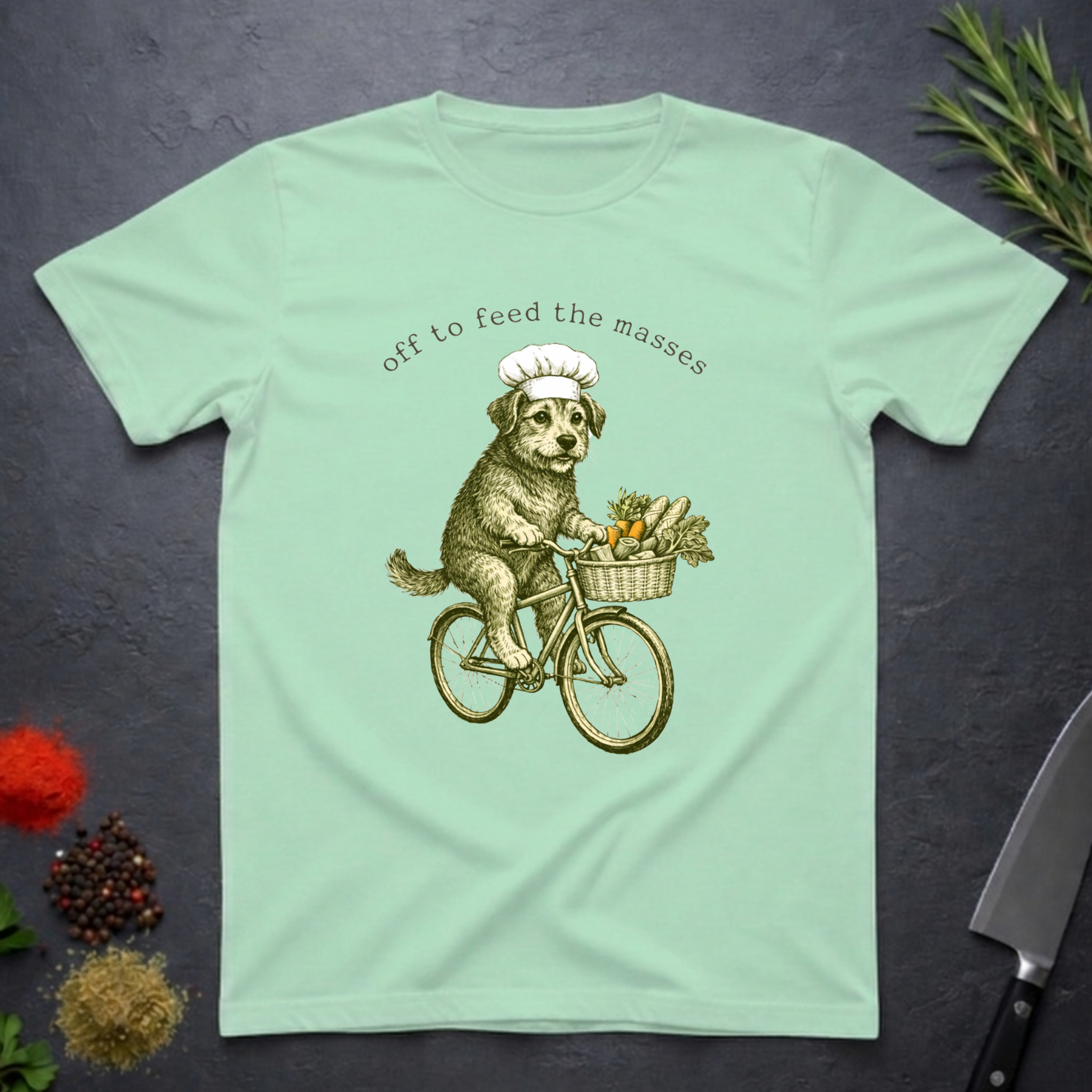 Mint green t-shirt with a graphic of a dog on a bicycle carrying a basket of vegetables and text 'off to feed the masses' on a dark surface.