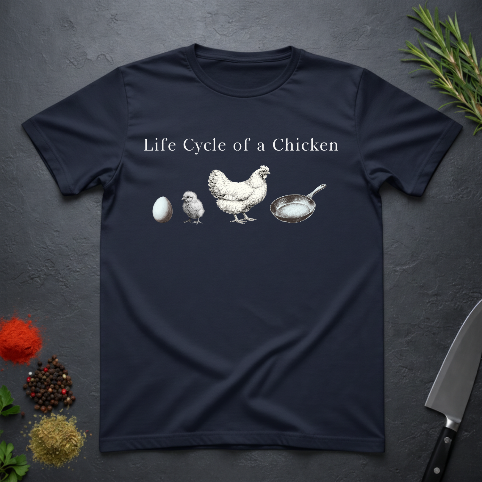 Navy blue t-shirt with text 'Life Cycle of a Chicken' and graphic of an egg, chick, hen, frying pan on a dark surface.