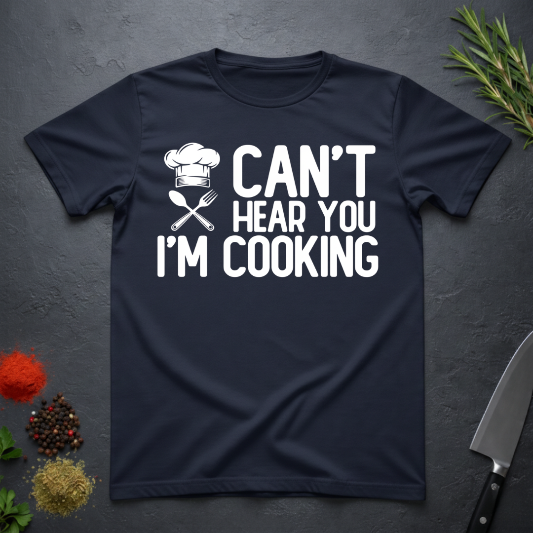 Navy blue t-shirt with 'Can't Hear You I'm Cooking' text on a dark surface with kitchen items.