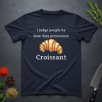 Navy blue t-shirt with a croissant graphic and text "I judge people by ho they pronounce Croissant" on a dark surface.