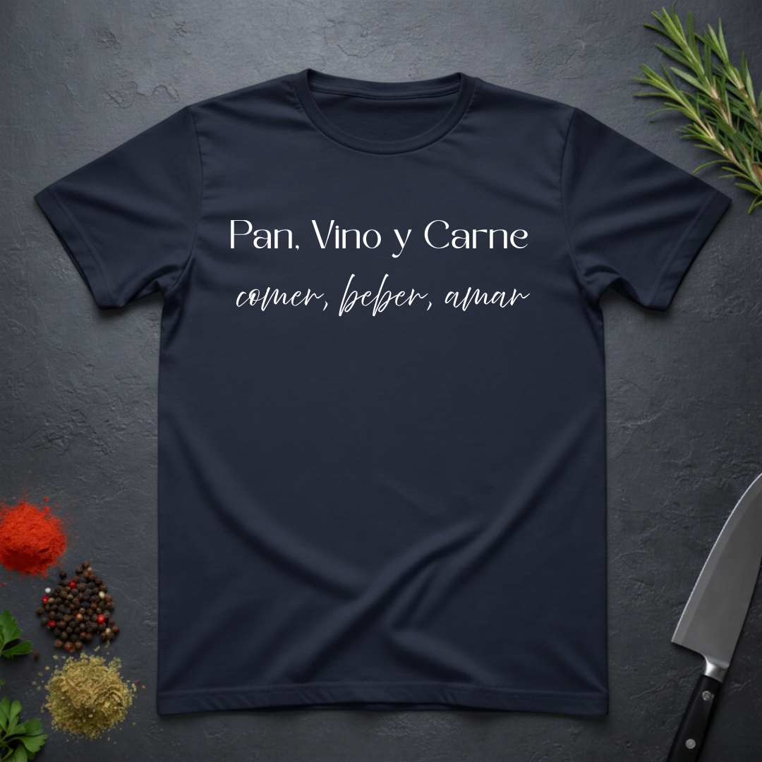 Navy Blue t-shirt with Spanish text "Pan, Vino y Carne" on a dark surface with decorative elements. 