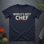 Navy blue t-shirt with 'World's Best Chef - and Dad' text on a dark surface with kitchen items.