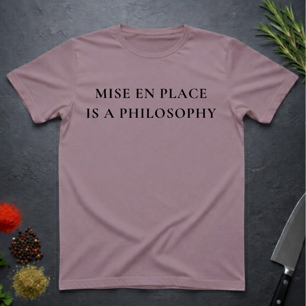 Paragon colored t-shirt with 'MISE EN PLACE IS A PHILOSOPHY' text on a dark surface with kitchen items.