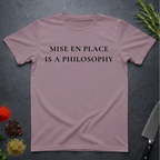 Paragon colored t-shirt with 'MISE EN PLACE IS A PHILOSOPHY' text on a dark surface with kitchen items.