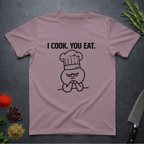 Paragon colored t-shirt with 'I COOK. YOU EAT.' text and graphic on a dark surface with kitchen items.