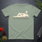 Sage Green t-shirt with a graphic of a chef lying on his side and text 'I Am the Secret Ingredient' on a dark surface with kitchen items.