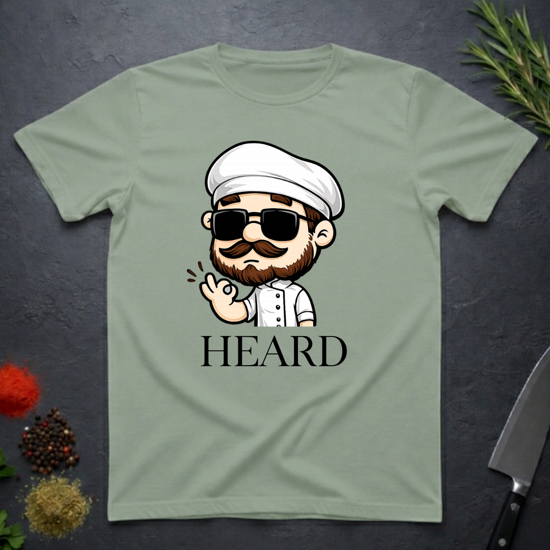 Sage green t-shirt with a cartoon character chef and 'HEARD' text on a dark surface.