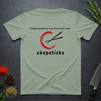 Sage green t-shirt with Chopsticks graphic and text "I judge people by how they hold their chopsticks" on a dark surface with kitchen items.